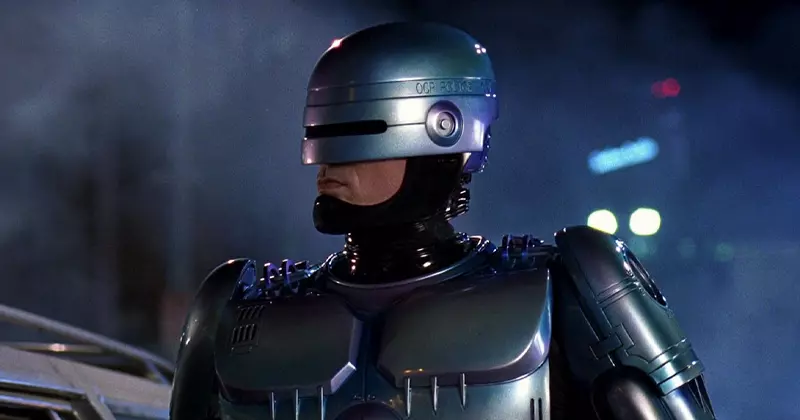 โรโบคอป หุ่นยนต์ตำรวจในชุดเกราะสีเงินและหมวกกันน็อคสีเงิน สวมไวเซอร์สีดำปิดบังใบหน้ามนุษย์ที่เหลืออยู่ เขากำลังมองออกไปจากกรอบภาพด้วยท่าทางแน่วแน่และดุดัน จากเรื่อง RoboCop | โรโบคอป (1987)